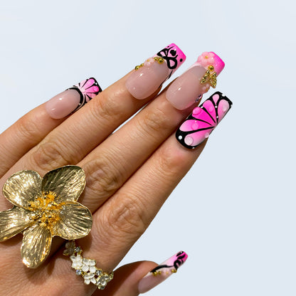 Hand wearing Sugar Noir Butterfly square nails styled with a large gold flower ring