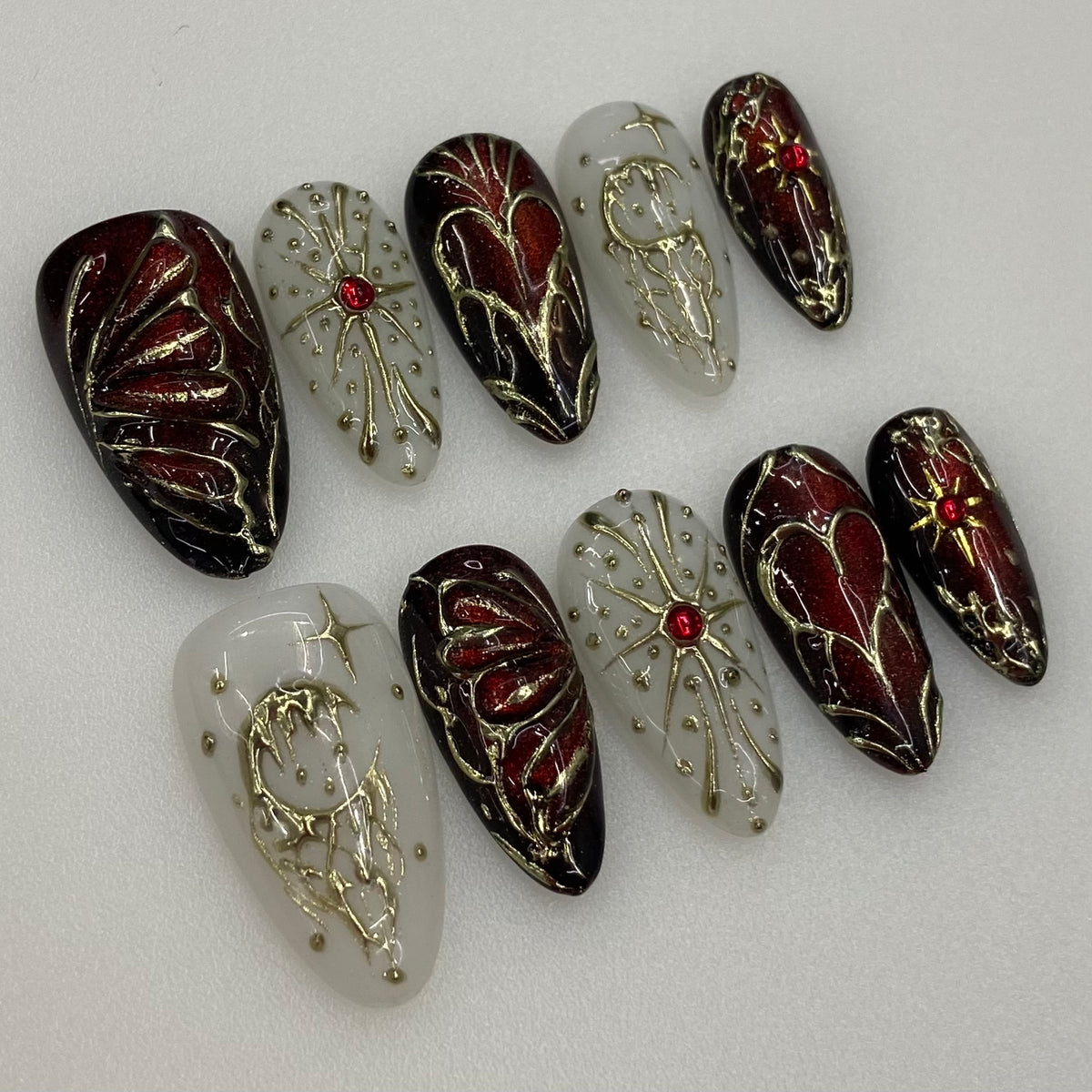 Close-up real photo of Crimson Alchemy press-on nails showing 3D gilded butterfly wings, ruby crystal starburst, and salon gel shine