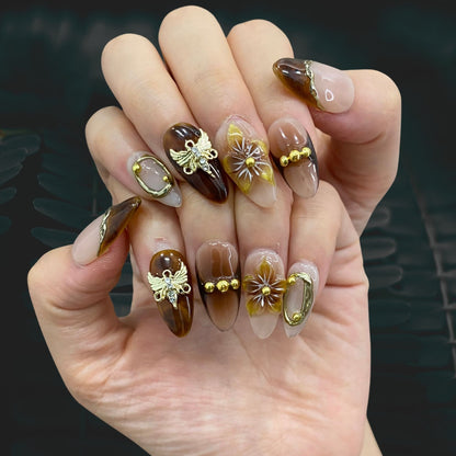 On-hand try-on of Gilded Terra nails showing 3D butterfly charms and gold bangles