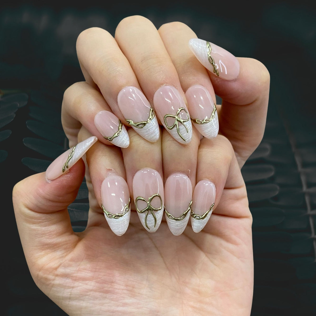 Both hands wearing Gilded Pearl Bow metallic gold ribbon nails in soft light